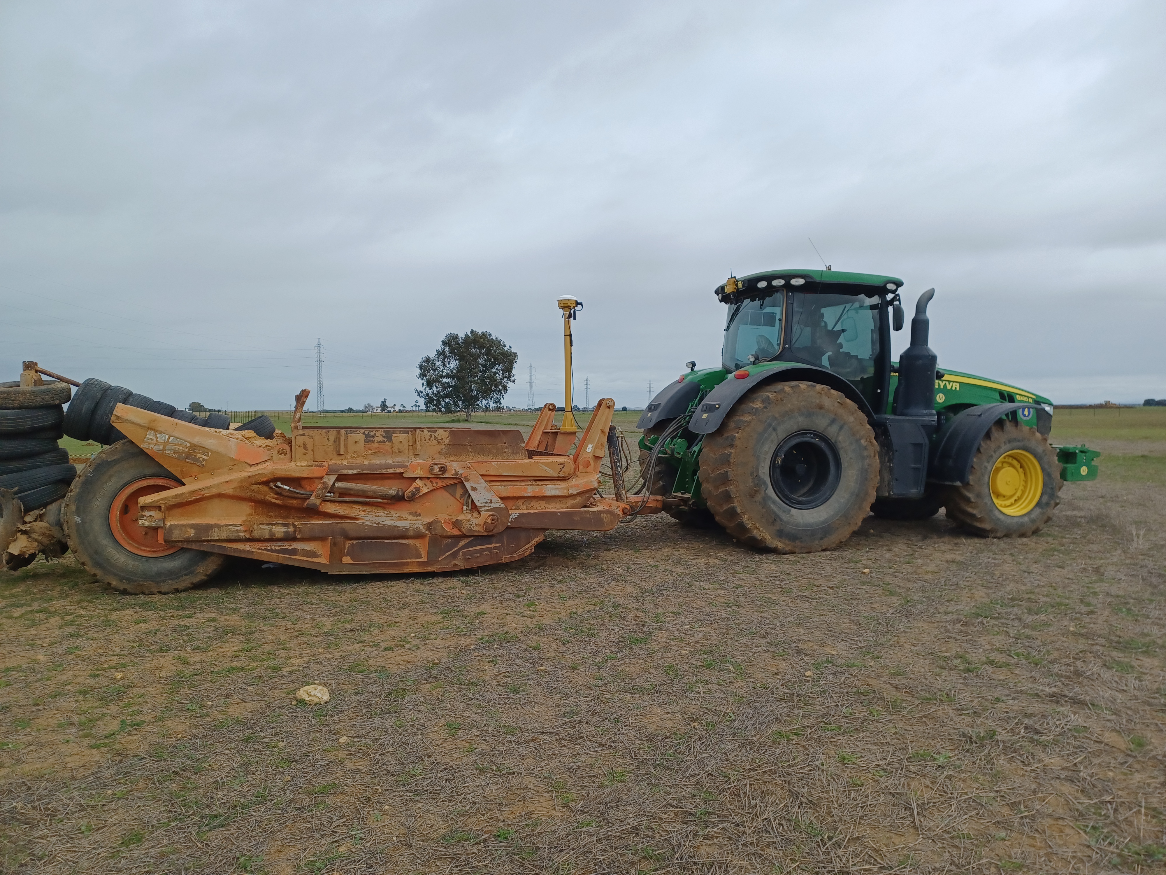 Trator agrícola John Deere equipado com o sistema 3D Trimble Earthworks, mostrando um implemento de nivelamento de terrenos em campo aberto. A avançada tecnologia de orientação GPS é visível na parte superior do trator, realçando a precisão e a eficiência nos trabalhos de construção