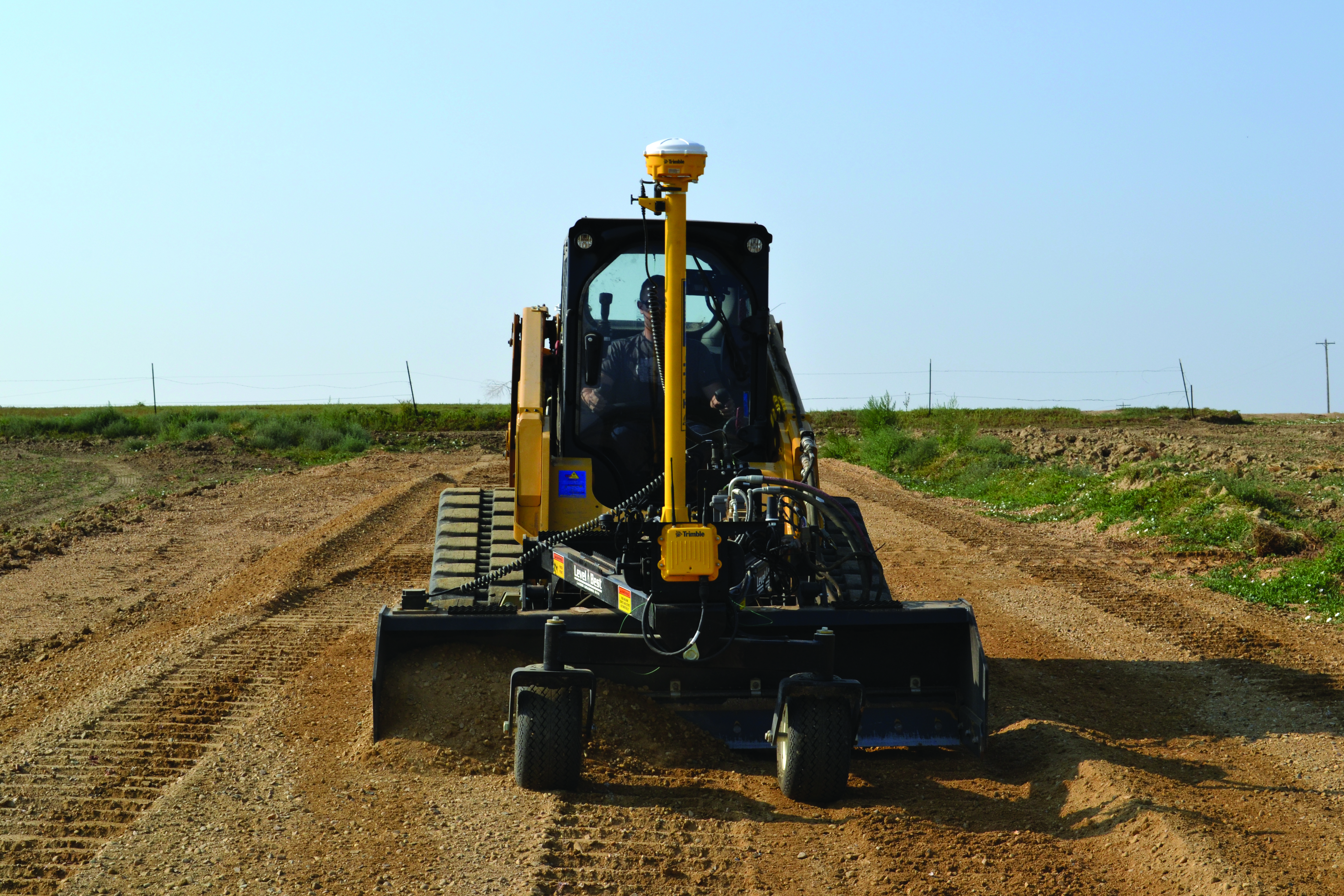 O sistema Trimble Earthworks 3D presente numa pá carregadora compacta Bobcat com um acessório de nivelamento num ambiente de construção. A avançada tecnologia GPS nos postes amarelos é visível, indicando uma maior capacidade de precisão para uma maior produtividade e eficiência na preparação do terreno