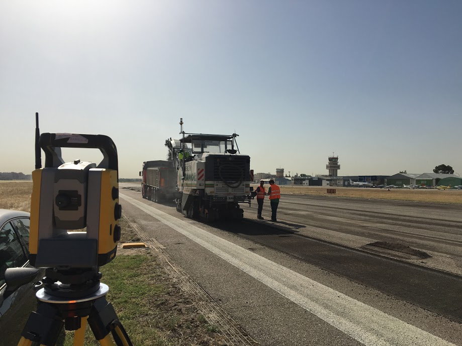 Estación total topográfica en primer plano utilizada en sistemas de guiado para fresadoras en obras viales. Operarios trabajando con maquinaria de pavimentación en una carretera en construcción